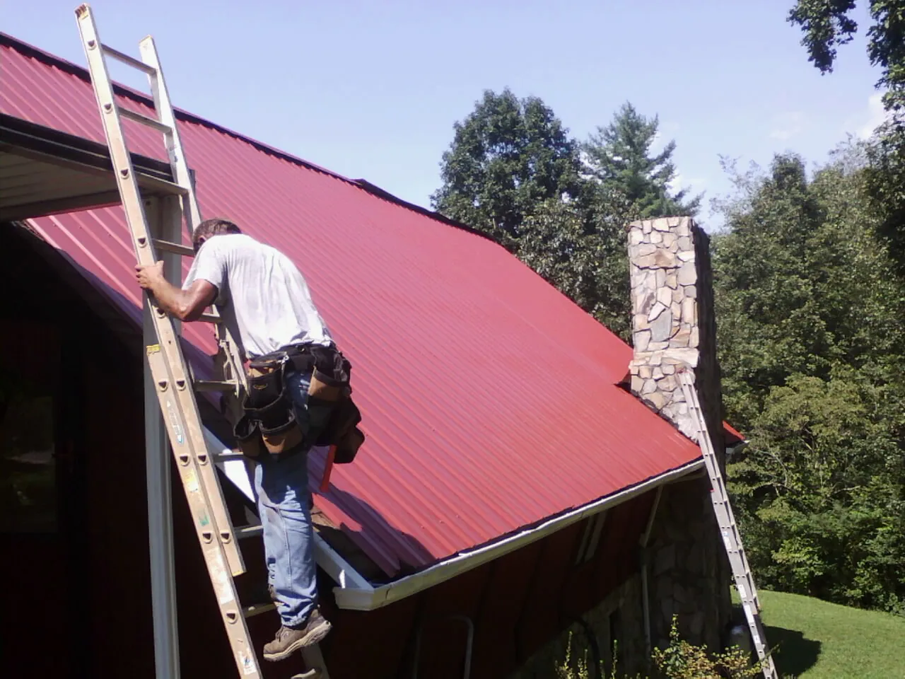 Roofer on a ladder beside a red metal roof and stone chimney — Soffit & Fascia Repair by Roofing Brothers in Andersonville